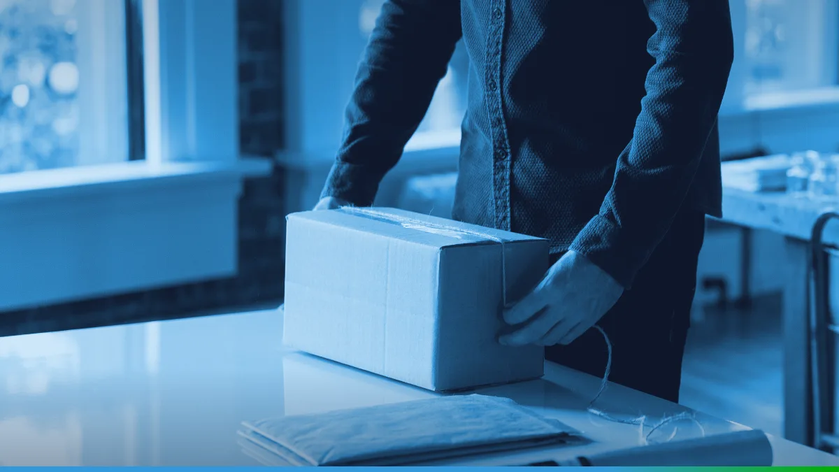 Pessoa em pé fechando uma caixa de papelão sobre uma mesa branca, perto de uma janela.