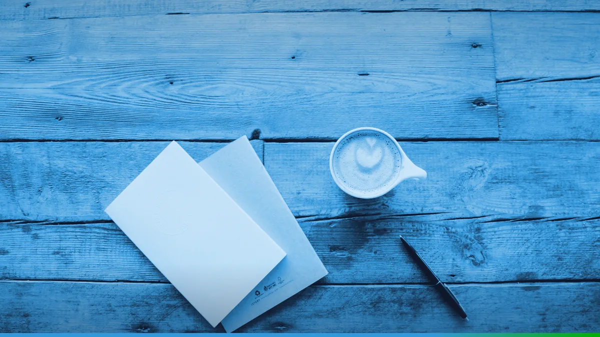 PW Host - Hospedagem de Sites Vitalícia e Sem Mensalidade Dois cadernos fechados, uma xícara de café com leite e uma caneta sobre uma mesa de madeira, vistos de cima.