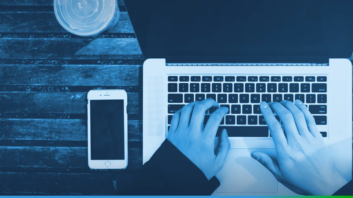 PW Host - Hospedagem de Sites Vitalícia e Sem Mensalidade Mãos digitando em um notebook visto de cima, sobre uma mesa de madeira, com um celular ao lado.