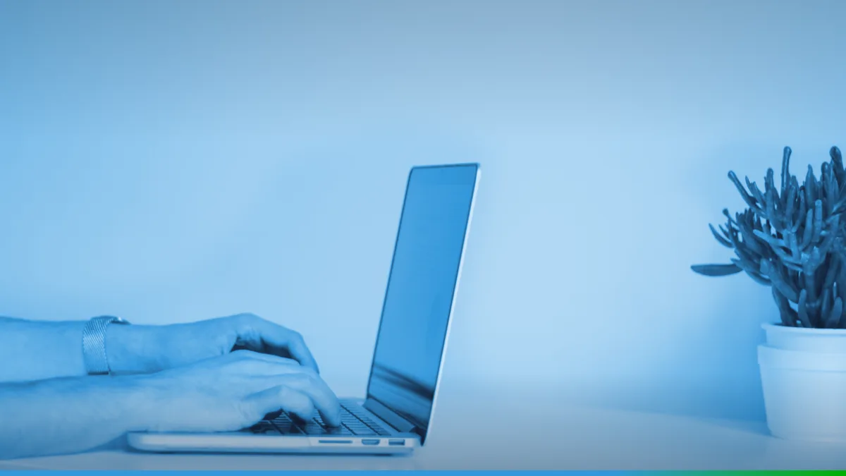 PW Host - Hospedagem de Sites Vitalícia e Sem Mensalidade Mãos digitando em um notebook prateado sobre uma mesa branca, com um vaso de planta à direita.